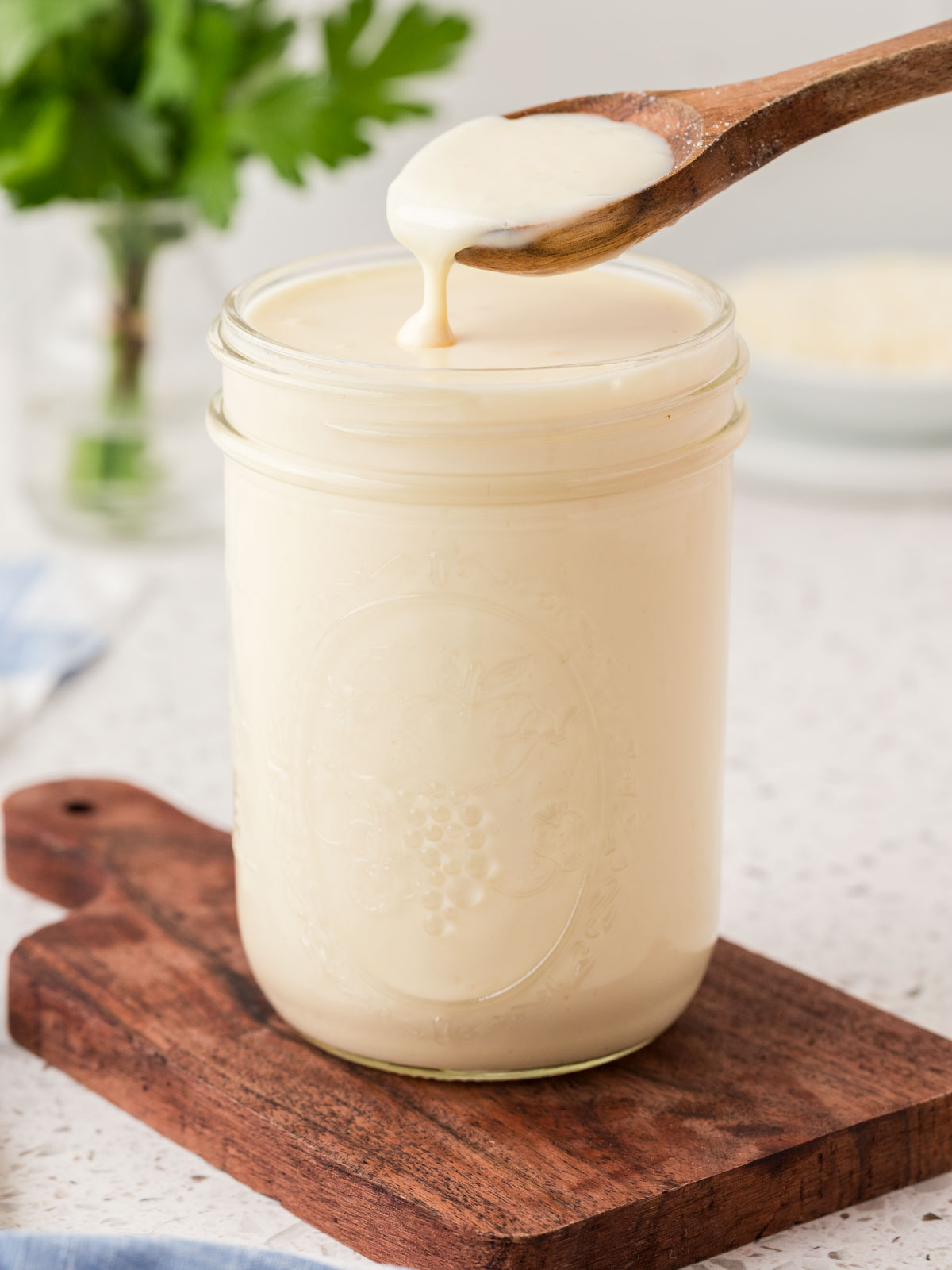 Homemade alfredo sauce in a jar sitting on a wooden board. A wooden spoon is dipping into the sauce. 