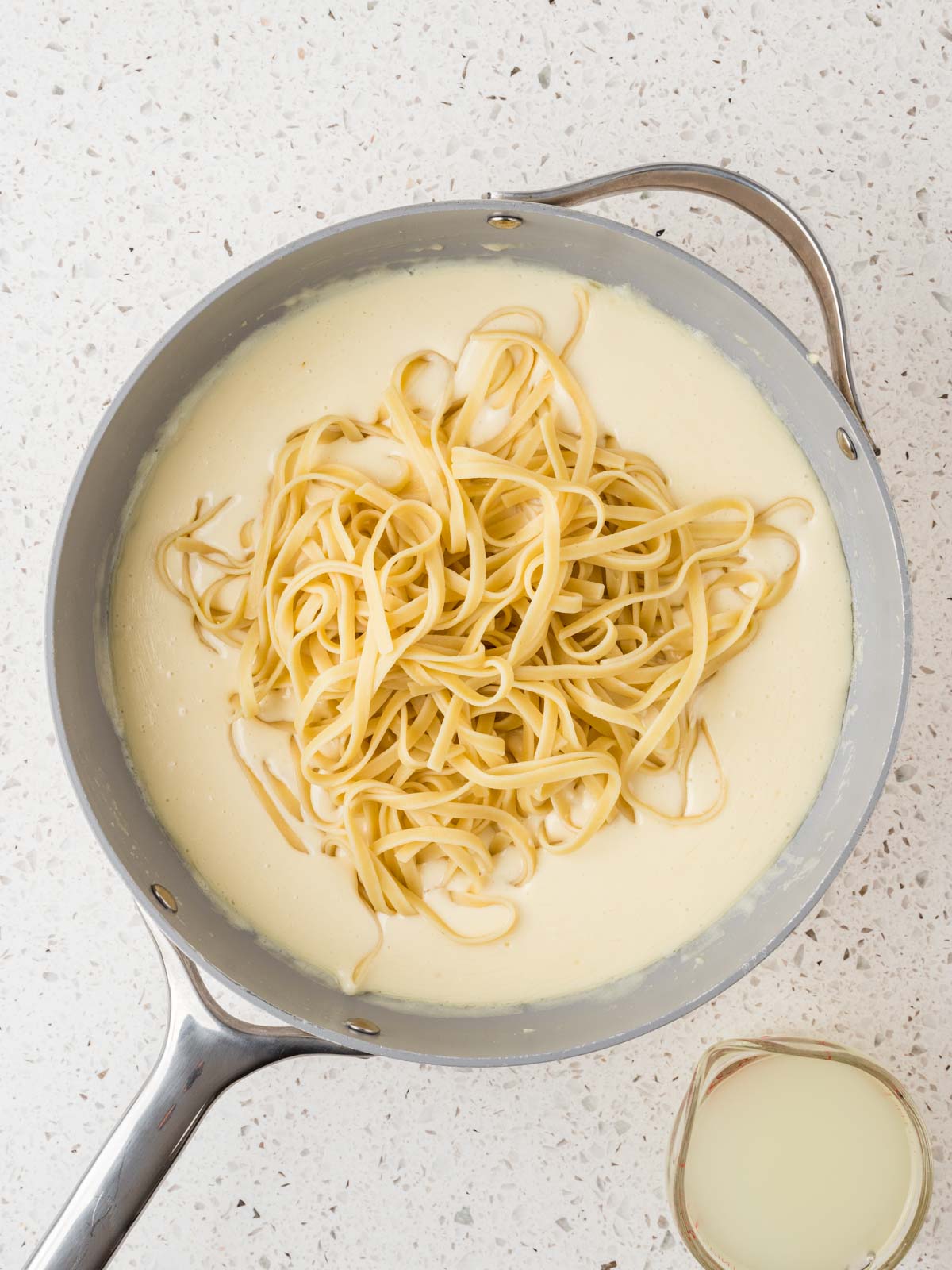 Cooked and drained fettuccine noodles in pan with alfredo sauce. Pasta water in a measuring cup set aside.