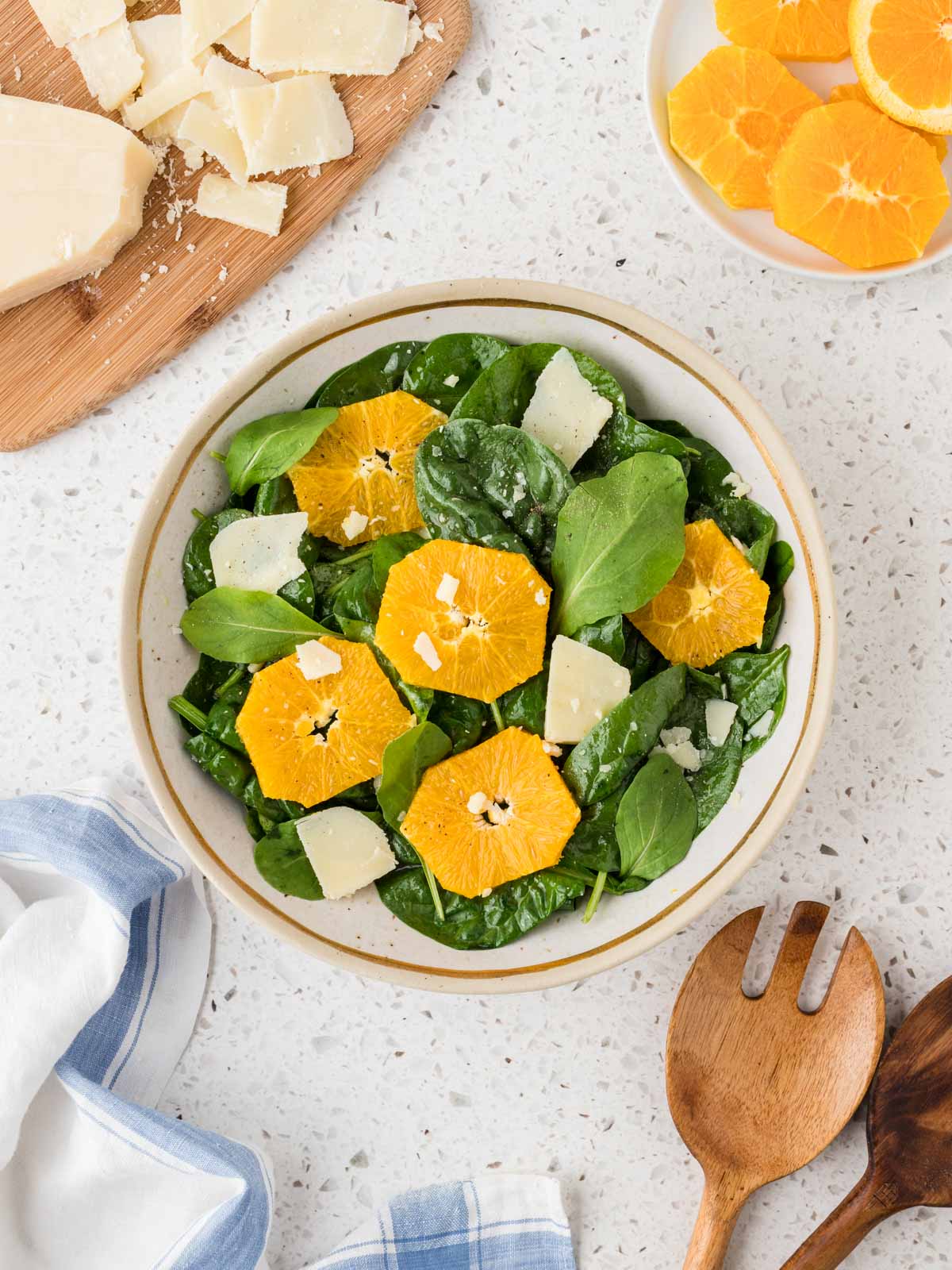 Arugula and Spinach Salad in a serving bowl surrounded by shaved parmesan cheese, sliced oranges and serving spoons. 