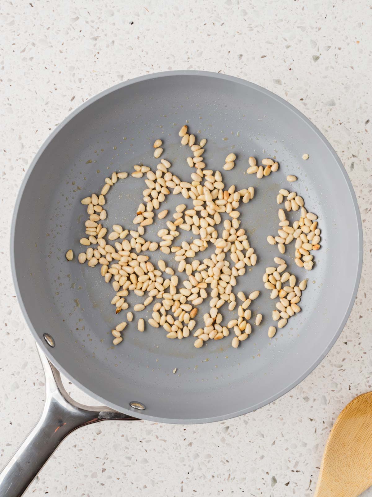 Pine nuts toasting a frying pan with a wooden spoon of to the side. 