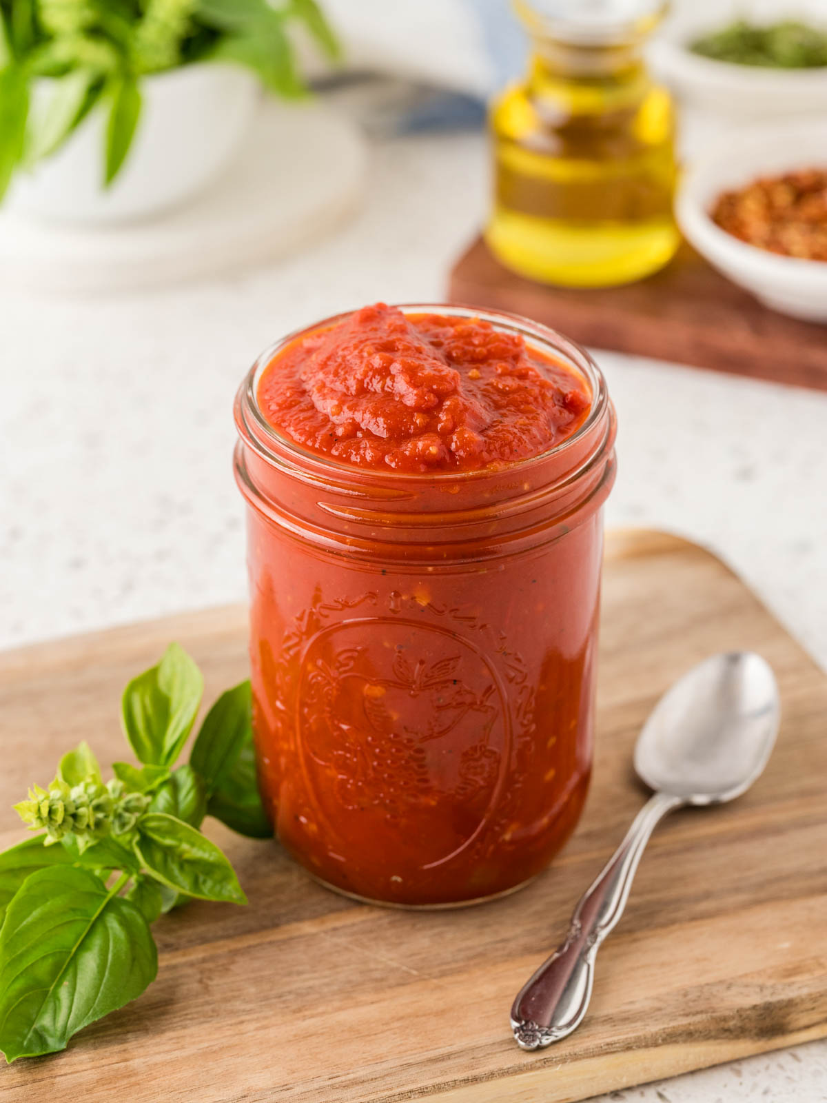Marinara sauce in a jar on a cutting board surrounded by fresh basil and a serving spoon.