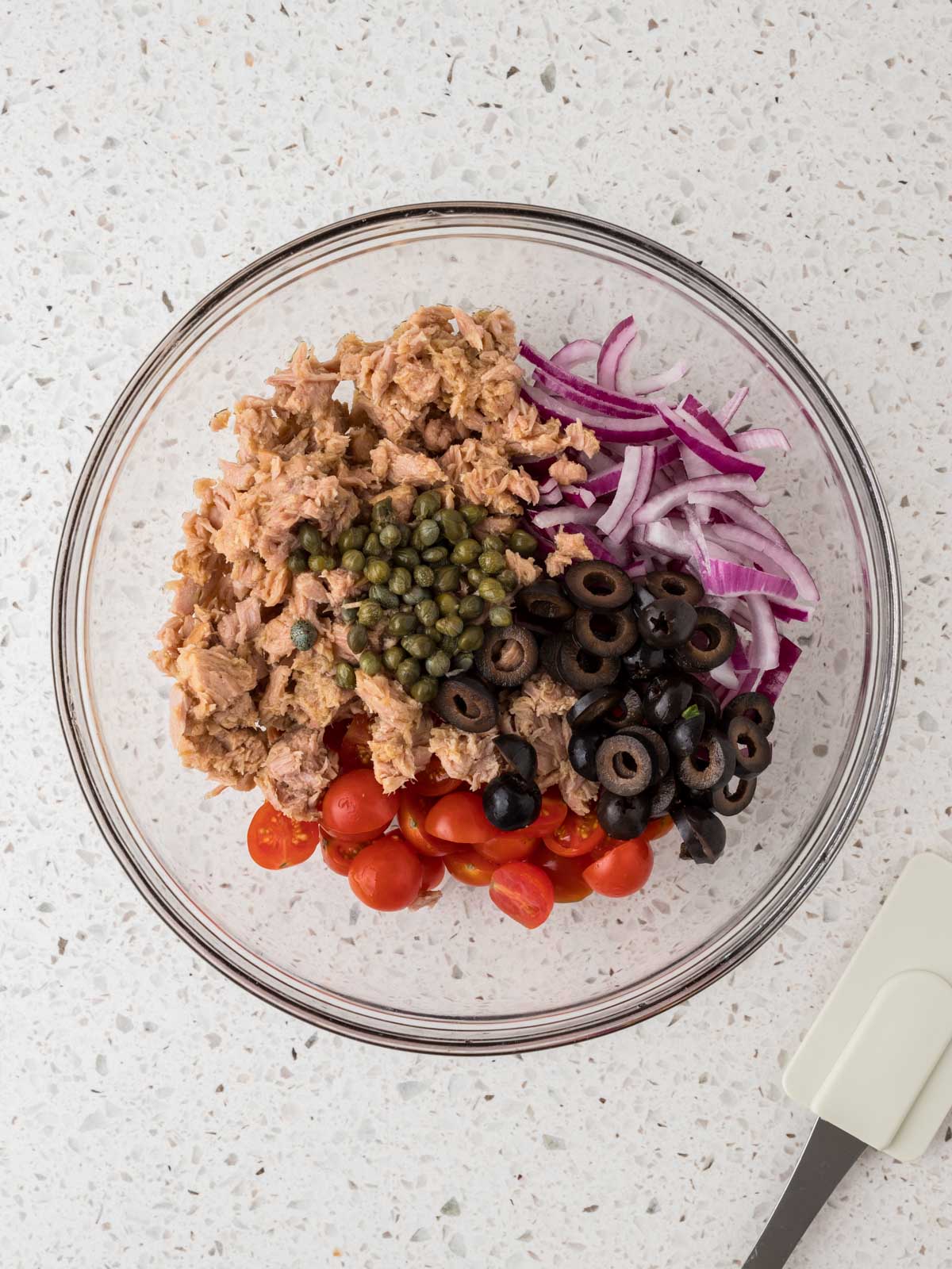 Glass mixing bowl with tuna, tomatoes, olives, capers, and red onions.