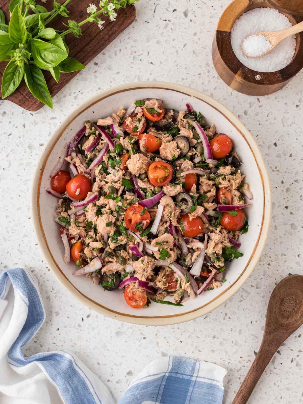 Italian Tuna Salad in a serving bowl surrounded by fresh basil, kosher salt and a wooden spoon. 