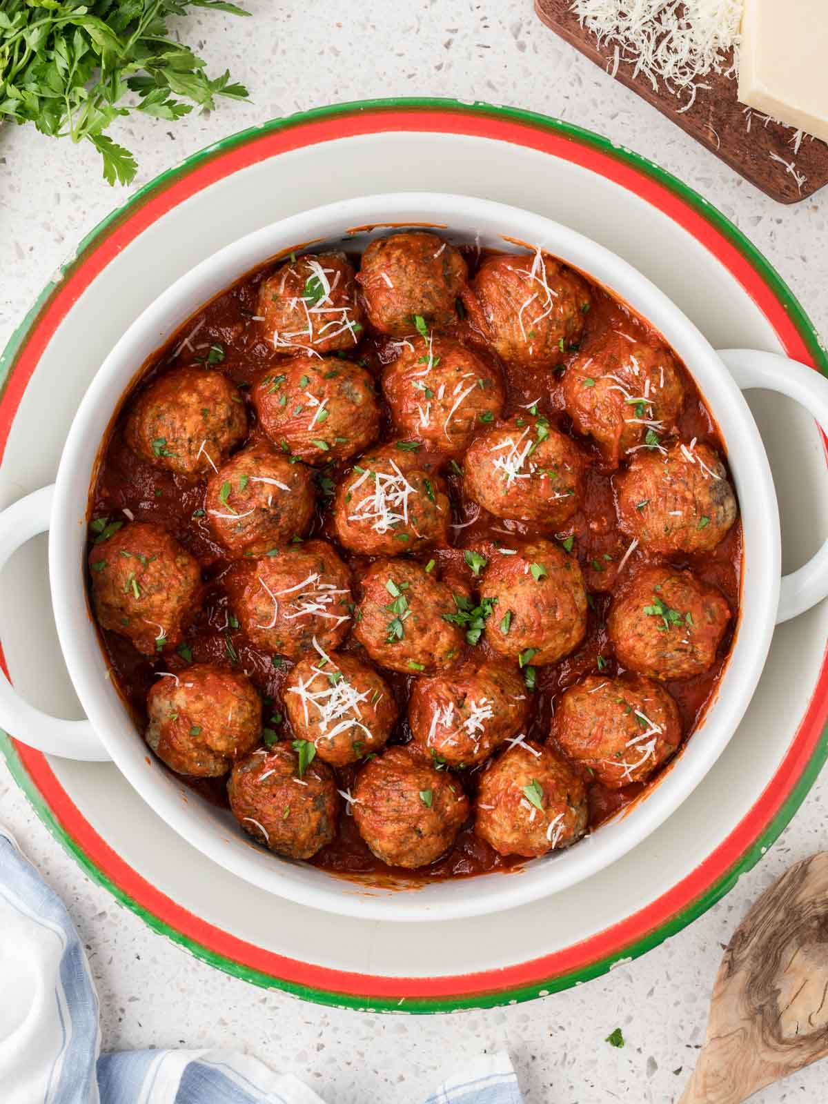 Homemade Italian Meatballs in a large serving bowl surrounded by Italian parsley, parmesan cheese and a serving spoon.