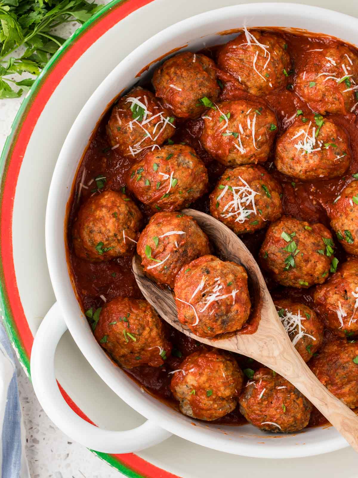 Homemade Italian Meatballs in a large serving bowl with 2 meatballs on a wooden spoon. Meatballs are garnished parmesan cheese and fresh chopped parsley.