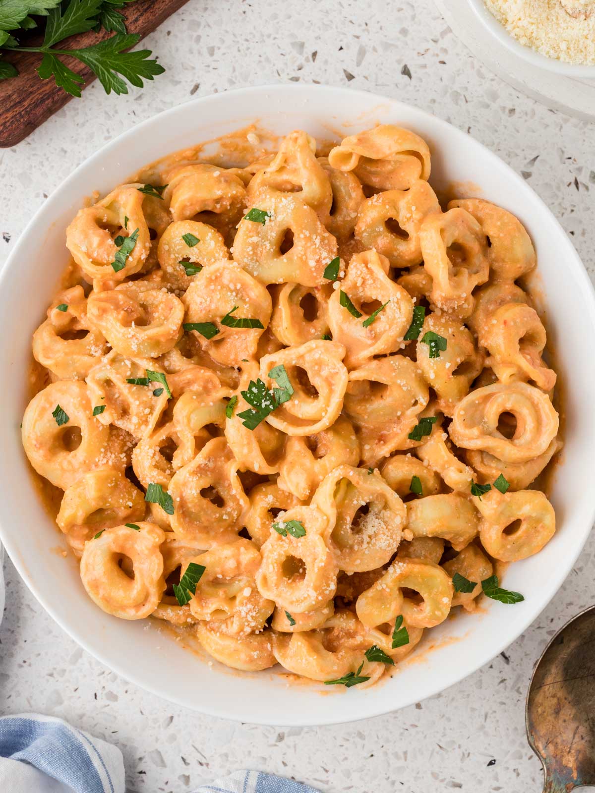 Creamy Tomato Tortellini in a serving bowl surrounded by parsley, parmesan and a serving spoon.