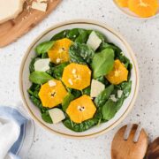 Arugula and spinach salad in a bowl surrounded by parmesan cheese, sliced oranges and wooden serving spoons.