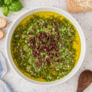 Basil pesto dipping oil in a serving bowl surrounded by fresh basil, parmesan cheese, and a wooden spoon.