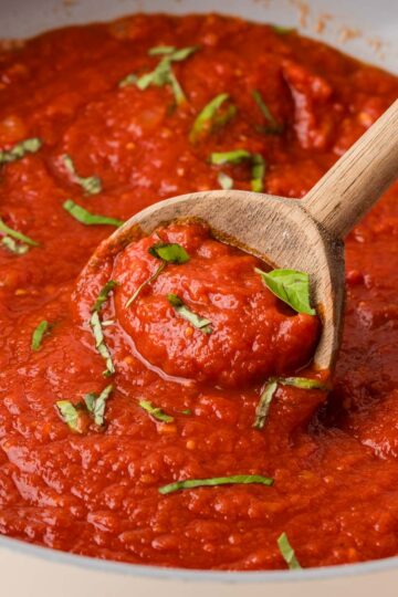 Easy marinara sauce prepared in a pan with a wooden spoonful of sauce. The marinara sauce is garnished with fresh basil.