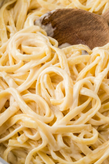 Homemade fettuccine alfredo prepared in a pan with a wooden spoon scooping the noodles.