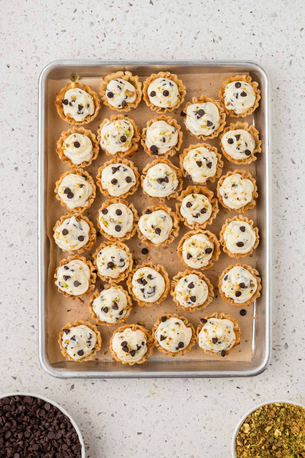 Cannoli bites on a cookie sheet garnished with mini chocolate chips and crushed pistachios.