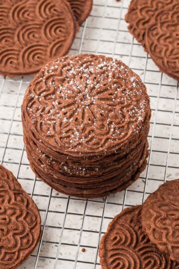 Chocolate pizzelles stacked on a cooling rack.